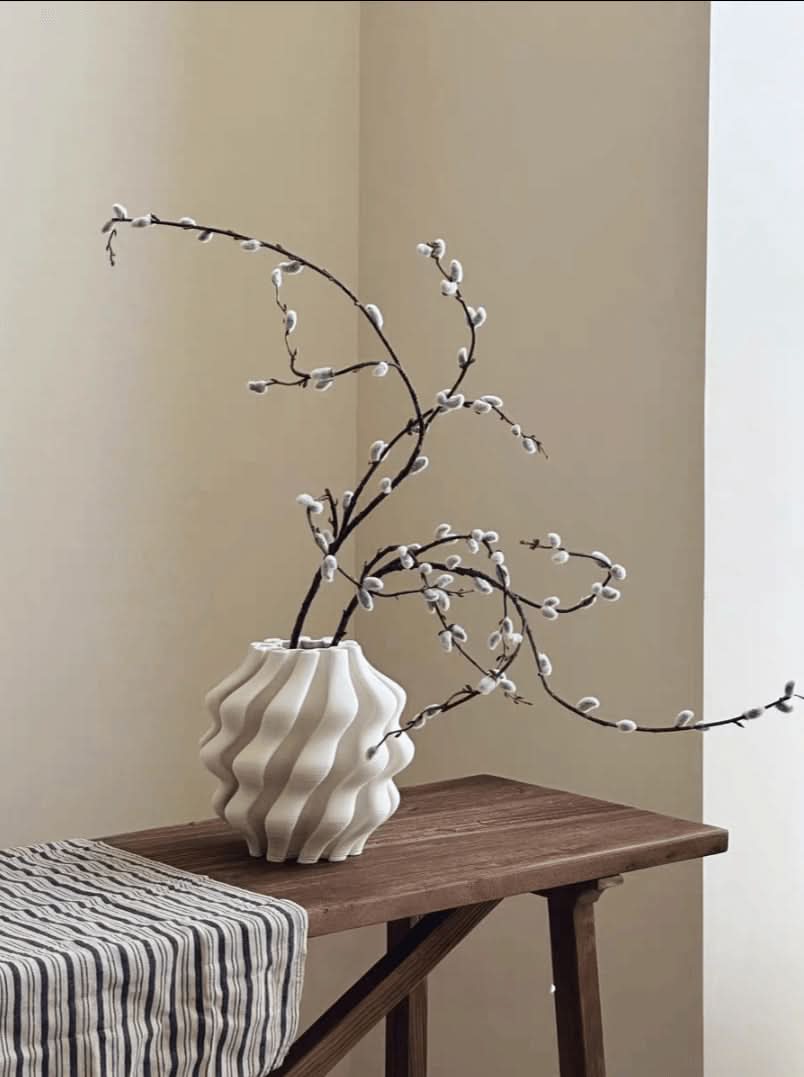 White 3D printed vase with wavy texture holds pussy willow branches on a wooden table.